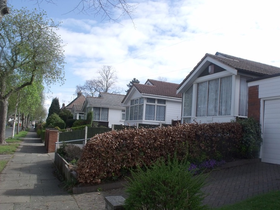 White timber clad bungalows behind hedges along a suburban street at the Austin Village in Northfield, Birmingham